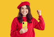 © Studio Romantic - Photo of beautiful brunette woman holding smartphone celebrating victory. Excited ecstatic girl wearing red beret and sweatshirt raising fist up in triumph saying yes on isolated yellow background