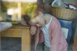 © PeterPike - Photo of a little tired girl in a pink blouse bowing her head while sitting at a wooden table behind a large glass window in her room. Difficult childhood concept