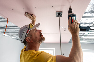 Wall Mural - The worker screwing plasterboard to the ceiling. He is using special electric screwdriver. A red plasterboard improve the fire resistance of ceiling structures.