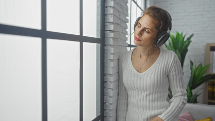 Wall Mural - A thoughtful young woman enjoys music with headphones in a cozy modern living room with plants and natural light.