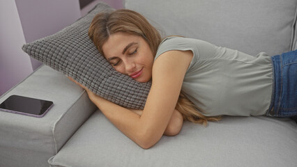 Poster - A beautiful young woman napping on a gray sofa with a smartphone nearby, indoors in a cozy living room setting.