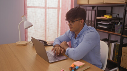 Wall Mural - Handsome hispanic man working on a laptop in an office setting with organized shelves and desk accessories.