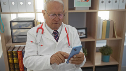 Sticker - Elderly caucasian man in a clinic room wearing a stethoscope and lab coat, using a smartphone, with bookshelves in the background.