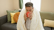© Krakenimages.com - Mature man enjoying coffee at home wrapped in a cozy blanket on a comfortable couch in a well-decorated living room