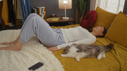 Poster - Young chinese woman relaxing on a bed in the bedroom with her pet cat nearby, while cozy indoor surroundings showcase a domestic and homely atmosphere.