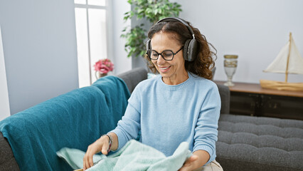 Sticker - A middle-aged hispanic woman with curly hair wearing glasses enjoys music through headphones while folding clothes in a cozy living room.