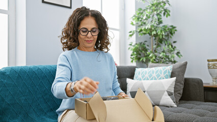 Canvas Print - Middle-aged woman with curly hair unpacking a box in a cozy living room setting, reflecting a serene domestic interior.