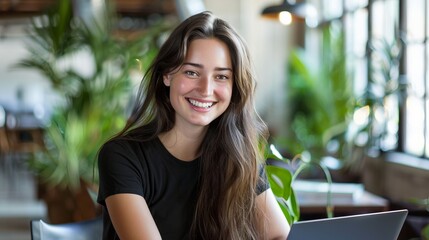 Poster - Young woman with long dark hair, smiling while working on a laptop in a modern, light-filled office space. 