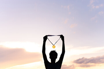 Silhouette of a male athlete raising his gold medal to celebrate his victory in a major sports event. In the background, there is a beautiful sky.