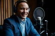 © sornram - A man in a suit and headphones sits in a recording studio, speaking into a professional microphone with a smile.