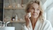 © Farid - Gorgeous mid age older adult 50 years old blonde woman wears bathrobe in bathroom applying nourishing antiage face skin care cream treatment, looking at mirror doing daily morning beauty routine.