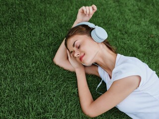 Wall Mural - Woman listening to music with headphones in a white T-shirt lying on the green grass of the lawn in the park, meditating with her eyes closed