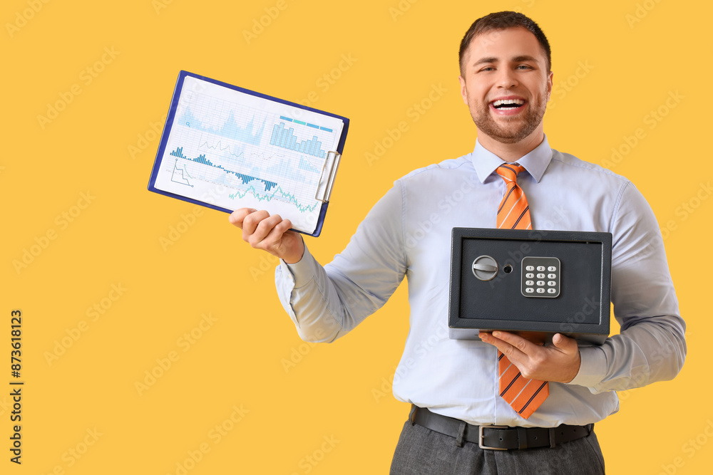 Young happy man holding safe box and clipboard with graphics on yellow background