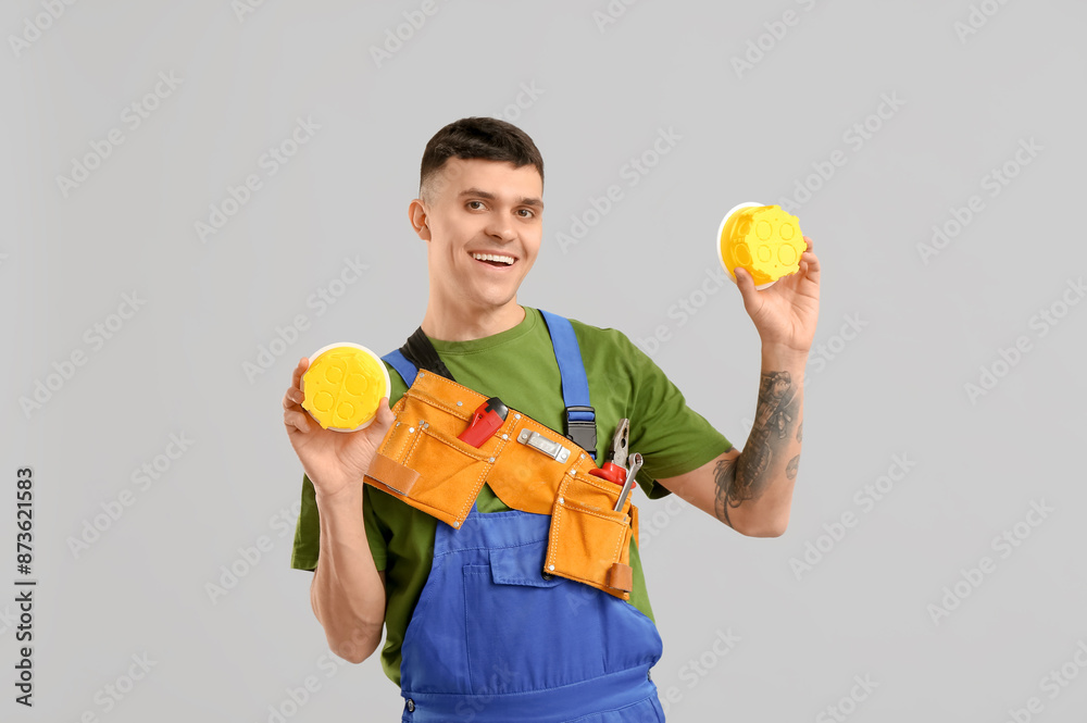 Young male electrician with tool bag and electrical junction boxes on grey background