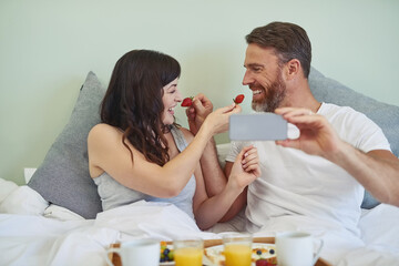 Canvas Print - Couple, relax and selfie with fruit in bedroom, bed and breakfast for rental on honeymoon vacation with love. Man, woman and together for feeding strawberry with photography, memory and commitment.