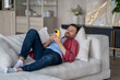 © DimaBerlin - Lazy man lies on sofa in living room, uses phone, thoughtlessly meaninglessly scrolling through feed of social networks, absorbing information, content. Tired calm guy resting on couch with smartphone