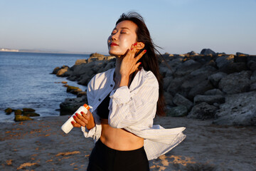 An Asian woman applying sunscreen to her face on the beach.  It is a bright sunny summer day. She is smiling as she rubs in the sunscreen.