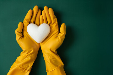 Fototapete - Female hands in yellow rubber gloves holding a white heart on a green background, from a top view. A cleaning service concept in the style of a valentine day or mother's Day banner template with space
