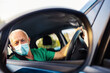 © Jelena Stanojkovic - Close up of a reflection in rear-view mirror of mature nurse driving a car, wearing face mask. Adult man driving to work wearing face mask during the day. Healthcare worker driving a car.