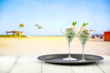 Wall Mural - Mint ice cream in glass cups on a tray on wooden table on a sandy tropical beach. Copy space for products.