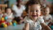 © familymedia - A cheerful toddler with curly hair, smiling brightly while looking directly at the camera, an indoor setting with other toddlers and colorful balls in the background.