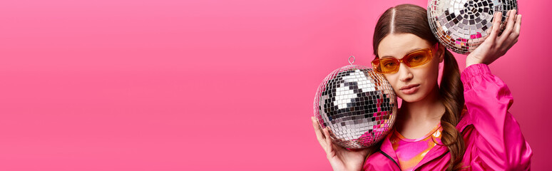 A young stylish woman in her 20s, wearing a pink shirt, joyfully holding two disco balls in a studio with a pink background.