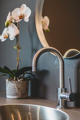 A close-up shot of the modern stainless steel faucet, which is positioned on one side of an elegant bathroom sink in front of a large mirror. A potted orchid sits next to it adding a touch of elegance