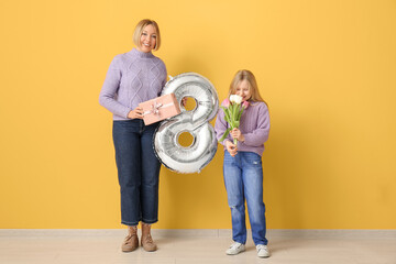 Sticker - Beautiful young woman with air balloon in shape of figure 8 and her cute little daughter holding bouquet of tulips near yellow wall