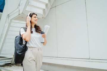 One young caucasian woman student going to university while listening to music on wireless headphones and holding papers and coffee	