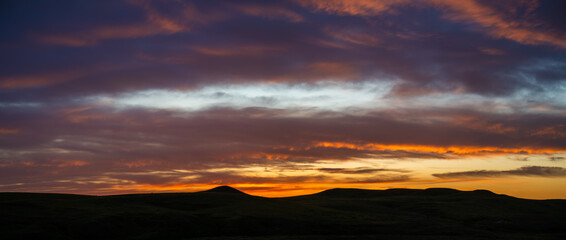 Wall Mural - Dramatic panoramic view of a multicolored sunset above a silhouette of rolling hills. The sky includes yellow, orange and light purple colored clouds.
