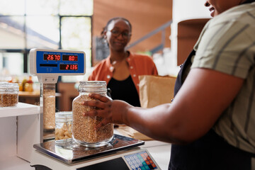 Wall Mural - African American woman in an apron assists a customer, weighing locally sourced grains on scale at fresh and organic grocery store. Local merchant measuring jar filled with bio food product.