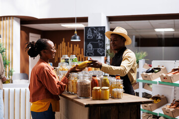 Canvas Print - African American male shopkeeper discussing with a female customer about different types of pasta sauce. Image shows young black woman receiving a bottle from vendor in local supermarket.