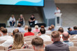 © Anton Gvozdikov - Large audience listens to speakers during a panel discussion at a tech conference. The room is filled with engaged attendees.
