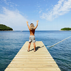 Wall Mural - Happy little boy with arms up, standing on wooden pier enjoying the sea- Travel, vacation, summer,freedom concept