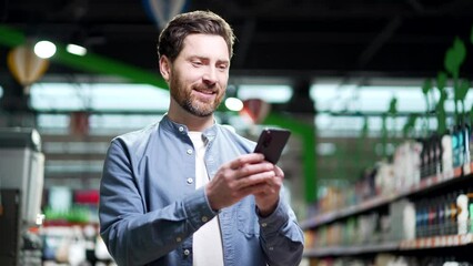 Happy adult man uses mobile phone in a supermarket while standing between the rows. Indoor Male typing text message, chatting browsing in smartphone in grocery store food market hypermarket or mall