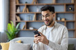 © Tetiana - Close-up photo of a happy young Indian man at home using a mobile phone, looking at the mobile screen in surprise