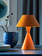 © SashaMagic - Modern orange table lamp next to blue ceramic vase on a desk.
