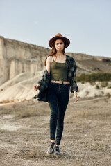 Wall Mural - stylish woman in plaid shirt and hat strolls confidently through the desert landscape with hands in pockets