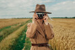 © Masson - Stylish Caucasian Blonde Woman in Brown Vintage Coat and Hat with retro camera in Wheat Field