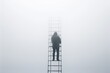 © Kishore Newton - A lone figure stands on a tall ladder, surrounded by thick fog. The minimalist scene evokes a sense of isolation and ascension.