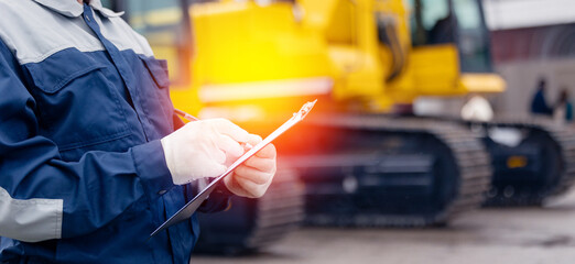 Wall Mural - Industrial worker male senior in helmet holds tablet computer on background of production of excavator factory.