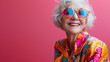 © Tijana - A joyful older woman with white hair wearing vibrant, colorful clothing and round sunglasses, smiling against a bright pink background.