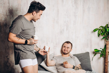 Wall Mural - Adult relationships. Two same-sex male partners resting together in one bed, having morning coffee. Romantic love. Homosexual couple