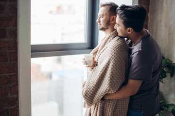 Wall Mural - Waist up portrait of homosexual man hugging his partner from behind, covered with soft plaid while standing at window holding hands