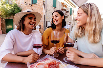 Young happy diverse women friends enjoying time together toasting red wine and eating italian aperitif in a restaurant. Friendship concept.