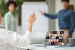 © Prostock-studio - Woman sitting in a meeting, raising her hand to ask a question. The room appears to be a hybrid setting, with the woman in person and other participants appearing on the laptop screen