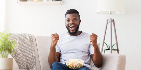 Wall Mural - Black man sits on a white couch, his arms raised in excitement, while he watches television. He is wearing a white t-shirt, blue jeans, and has a bowl of popcorn in his lap