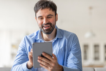 Poster - A man is smiling as he uses a tablet. He is wearing a light blue button-down shirt and is sitting indoors. He appears to be enjoying his time using the device.
