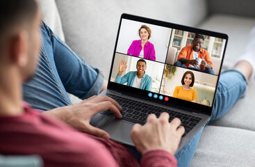 Poster - A man sits on a couch with his legs extended and uses a laptop computer to participate in a video conference call. The laptop screen displays four different video feeds of other people
