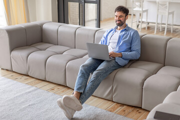 Poster - A man sits on a grey modular sofa in a modern living room, using a laptop computer. He is smiling and looks relaxed, possibly enjoying a break from work, high angle view